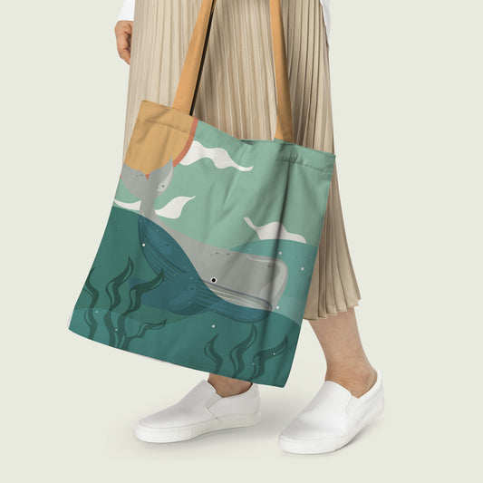 Women Carrying a tote bag with a print of a whale on the surface of the ocean where below is the sea with some corals and above is the sky with clouds and land.